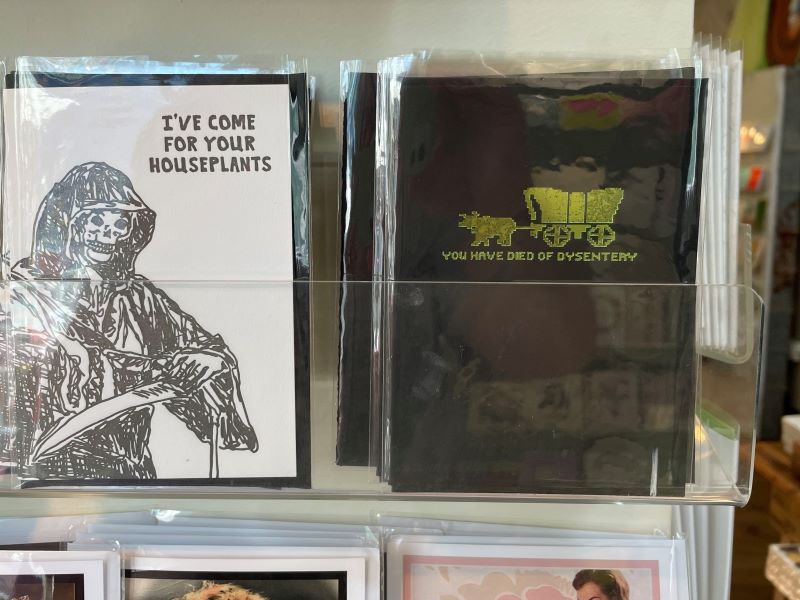 Novelty note cards from Oblation Papers: Card 1 features a grim reaper and says "I've come for your houseplants". Card 2 features an 8-bit graphic of a covered wagon pulled by an ox, and says "You have died of dysentery".